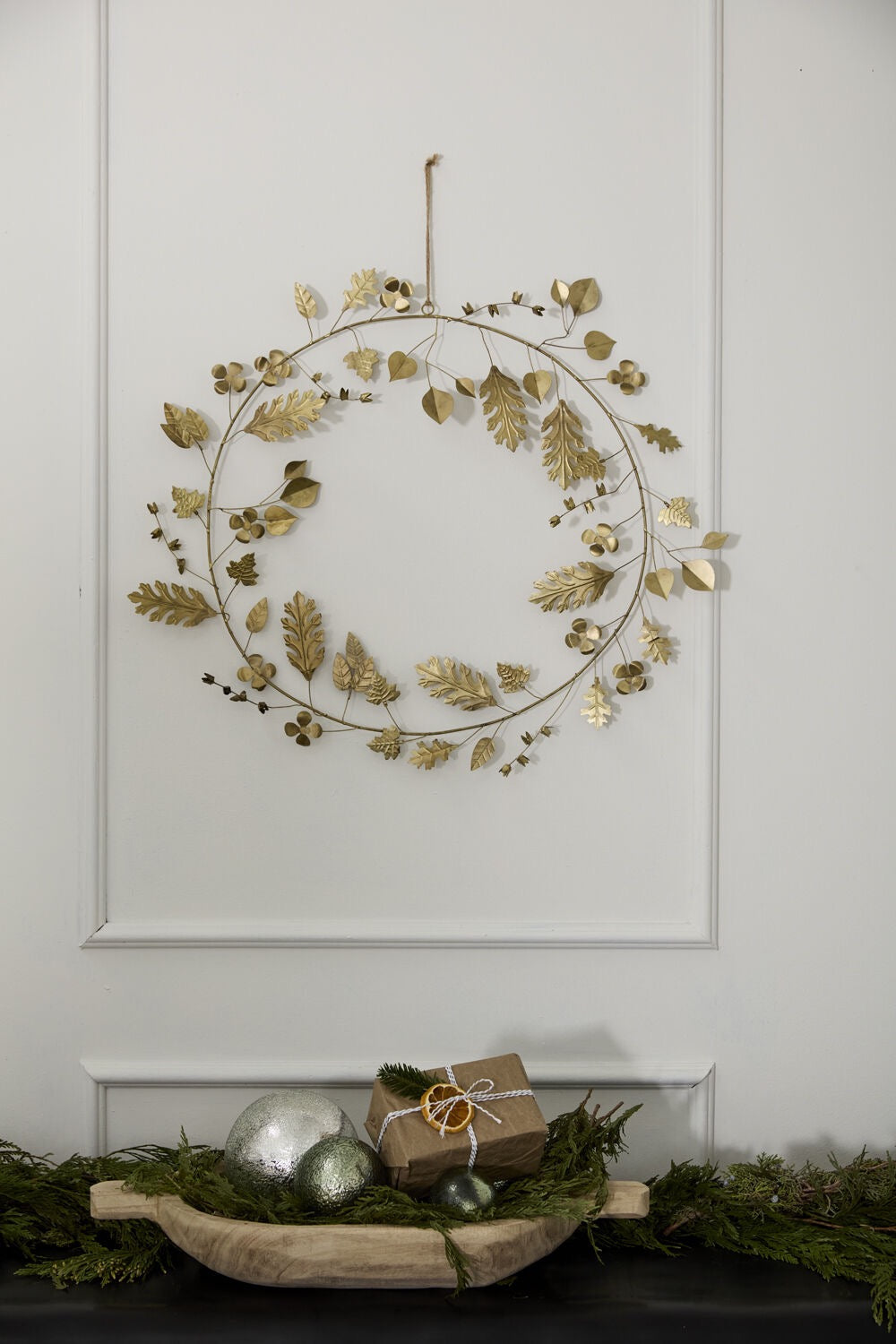 Decorative wreath with gold leaves on a white wall, with a small gift and greenery below.