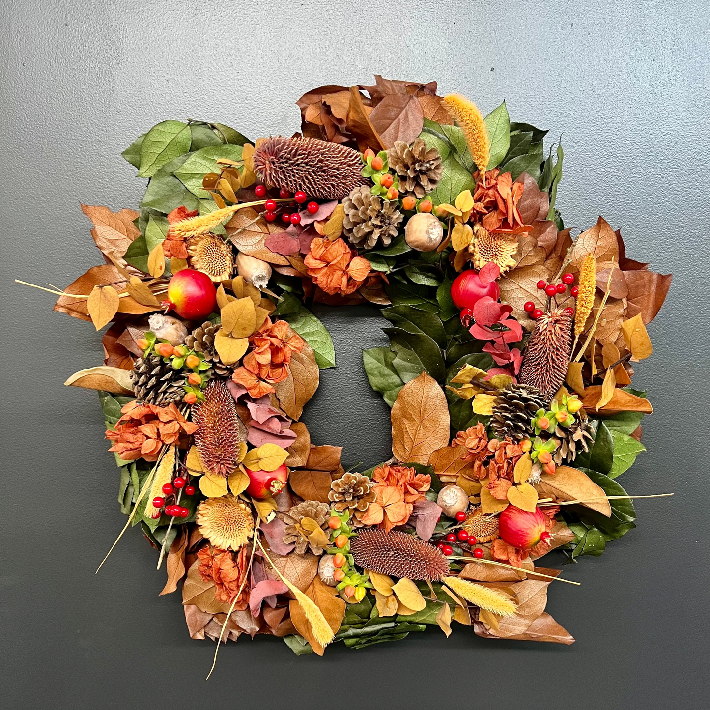 A handcrafted wreath made with various dried flowers and foliage, featuring rich oranges, yellows, reds, and burnished tones, and adorned with pinecones, berries, and a lemon leaf.