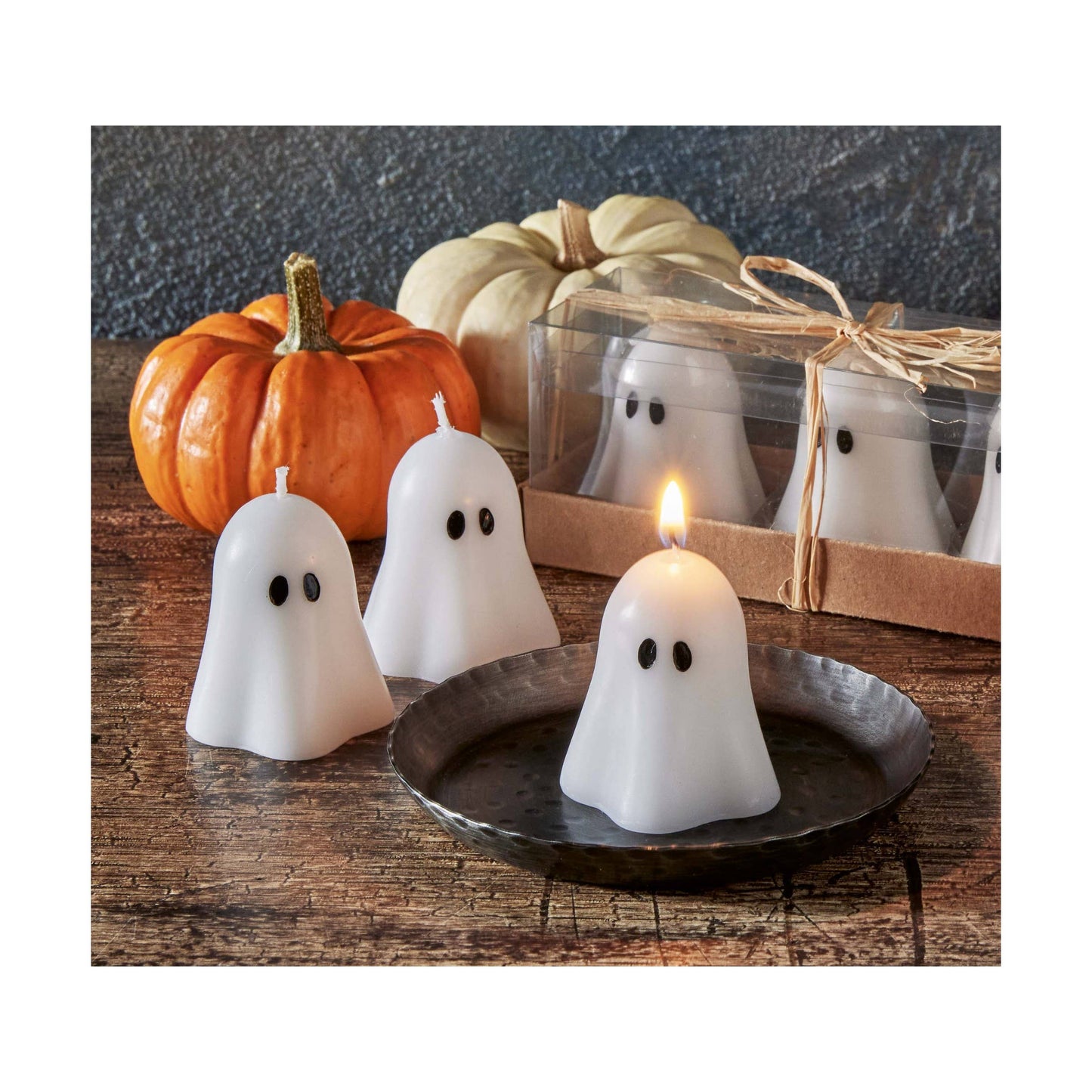 Ghost-shaped candles on a wooden surface with pumpkins and a box in the background.