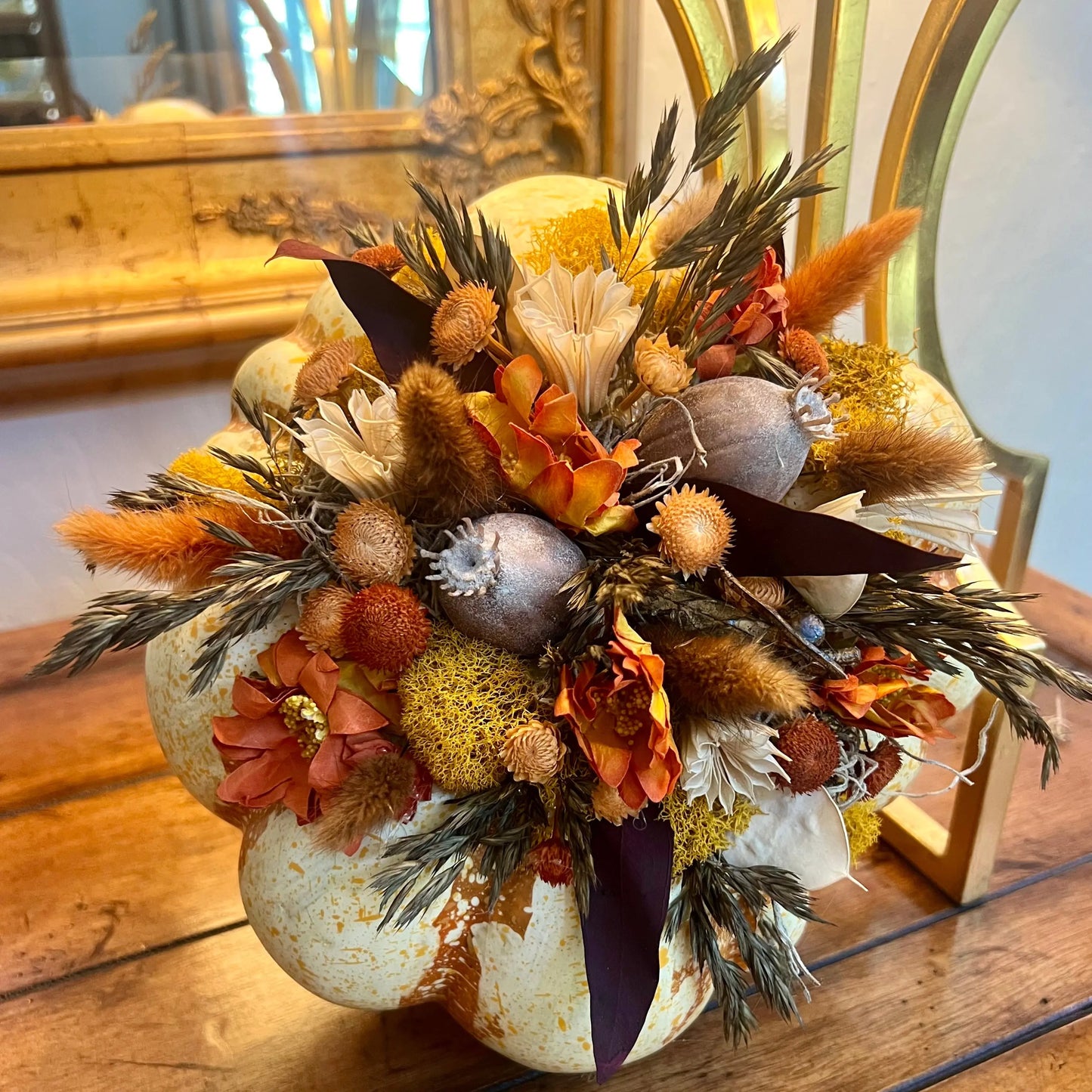 An autumn-themed floral arrangement featuring dried pumpkins, orange and yellow flowers, and natural brown grasses, displayed on a rustic wooden table.