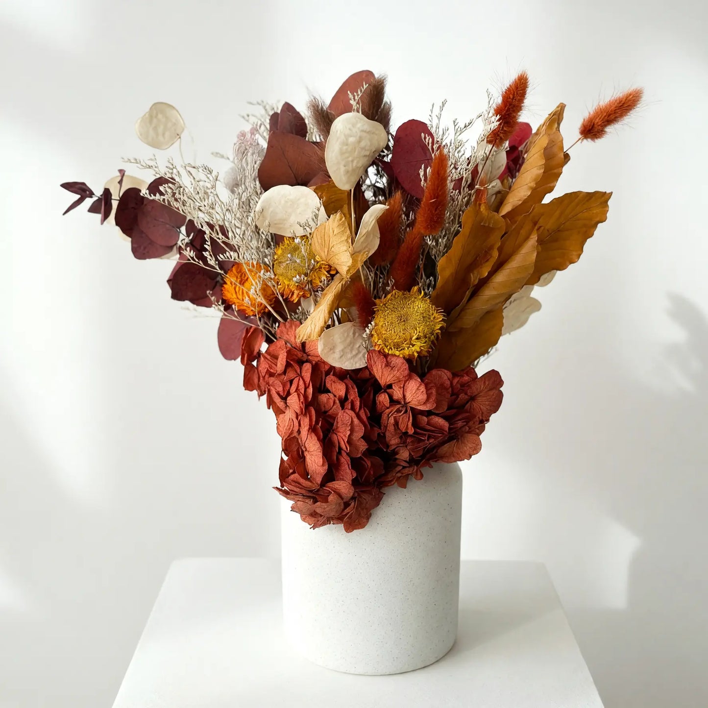 A dried flower arrangement in a matte white cylindrical vase, displayed on a white pedestal against a white wall with soft shadows.
