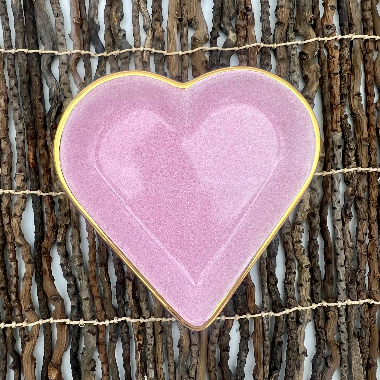 A heart-shaped porcelain trinket dish with a pink speckle glaze and hand-painted gold edge, displayed on a wicker surface.