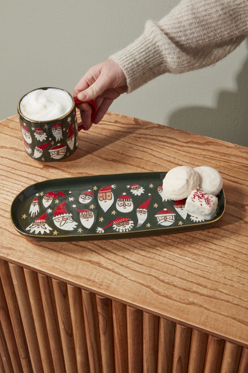 Person holding a mug with a decorative tray featuring Santa Claus designs on a wooden surface.