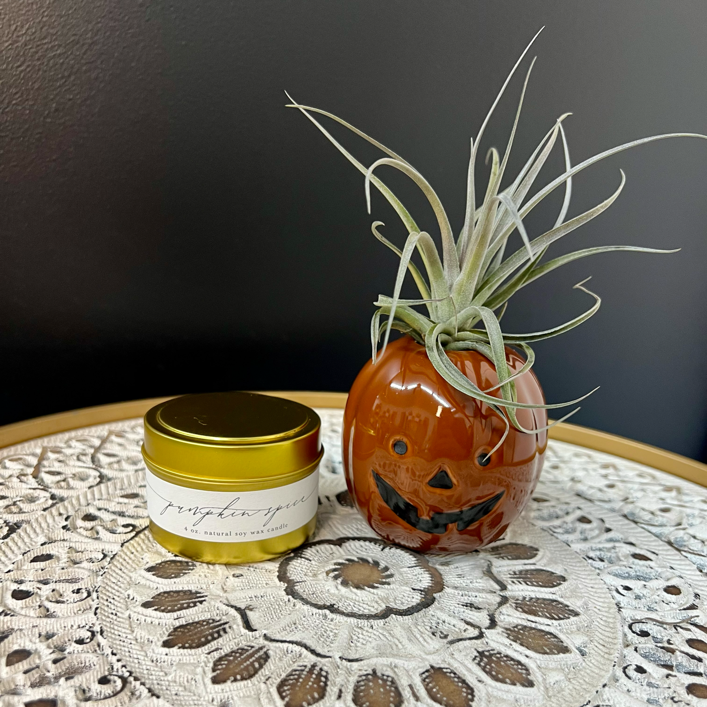 A ceramic pumpkin-shaped air plant holder with a live air plant on top, accompanied by a Pumpkin Spice candle in a gold jar.