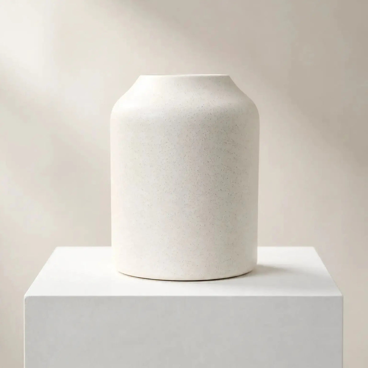 A matte off-white ceramic vase with a flared rim, displayed on a matching white pedestal against a warm beige background with soft diagonal shadows.