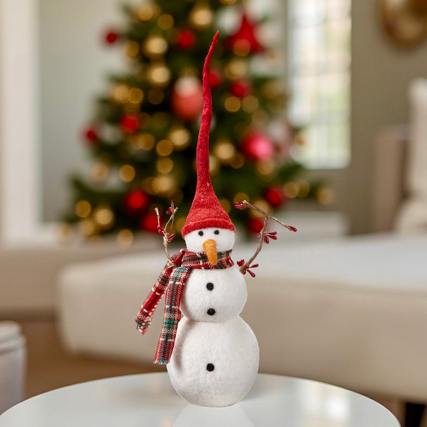 Decorative snowman with a red hat and plaid scarf on a table with a Christmas tree in the background.