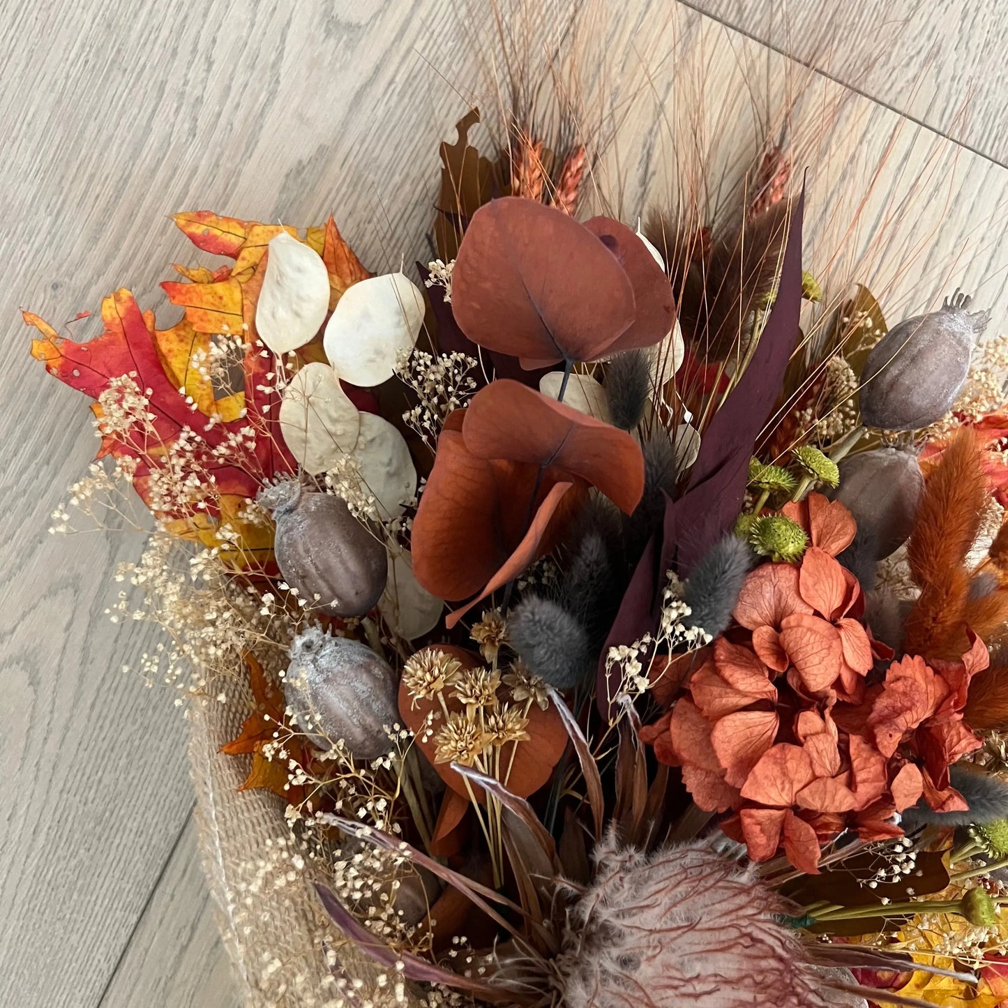 A dried autumn floral arrangement in a burlap-wrapped basket, featuring terracotta calla lilies, orange hydrangea-like blooms, beige baby's breath, and warm fall foliage on a light wood floor.