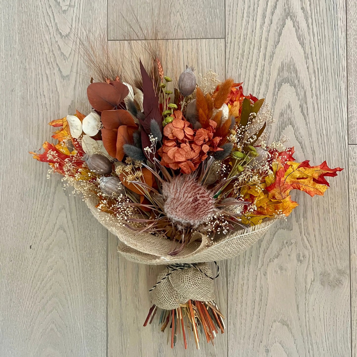 An autumn-themed dried flower bouquet wrapped in natural burlap, featuring orange, red, brown, and beige botanical elements, photographed from above on a light gray wood plank background.