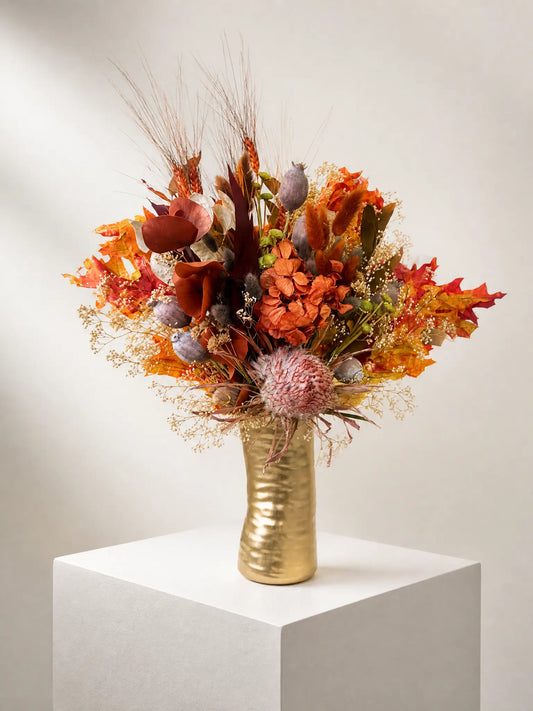 An autumn floral arrangement in a textured gold vase, displayed on a white pedestal against a neutral beige background.