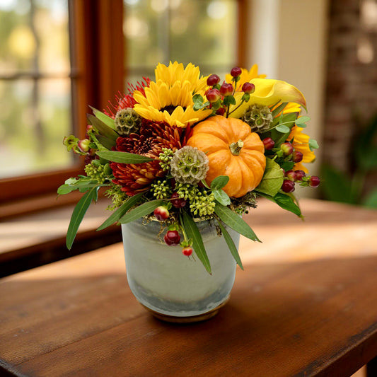 Autumn-themed floral arrangement with pumpkins and sunflowers in a vase on a wooden table
