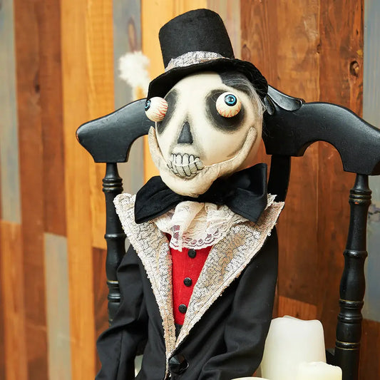 Joe Spencer Skeleton doll in a top hat and coat sitting on a chair with a wooden background