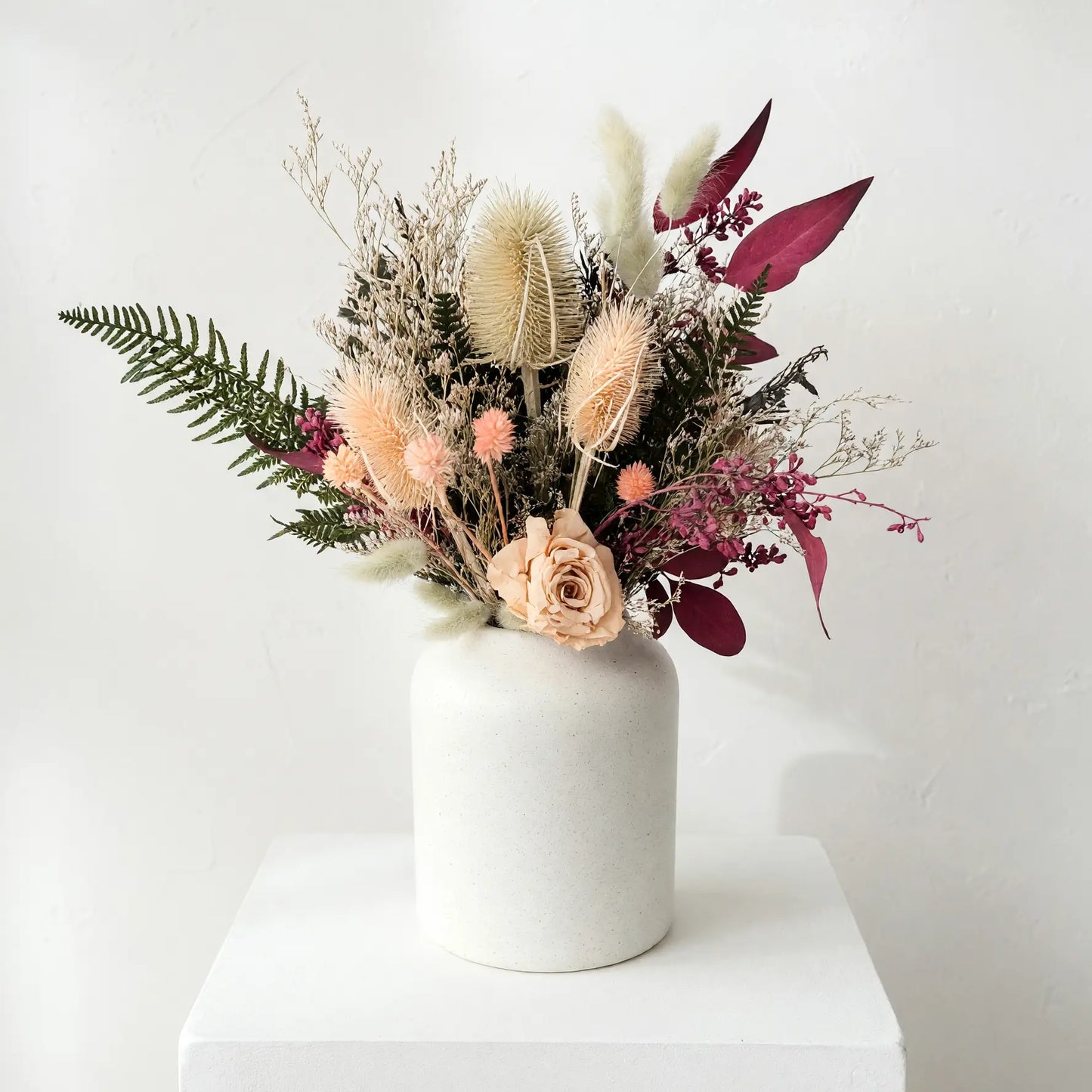 A dried flower arrangement in a matte white ceramic vase, displayed on a white pedestal against a light gray wall.