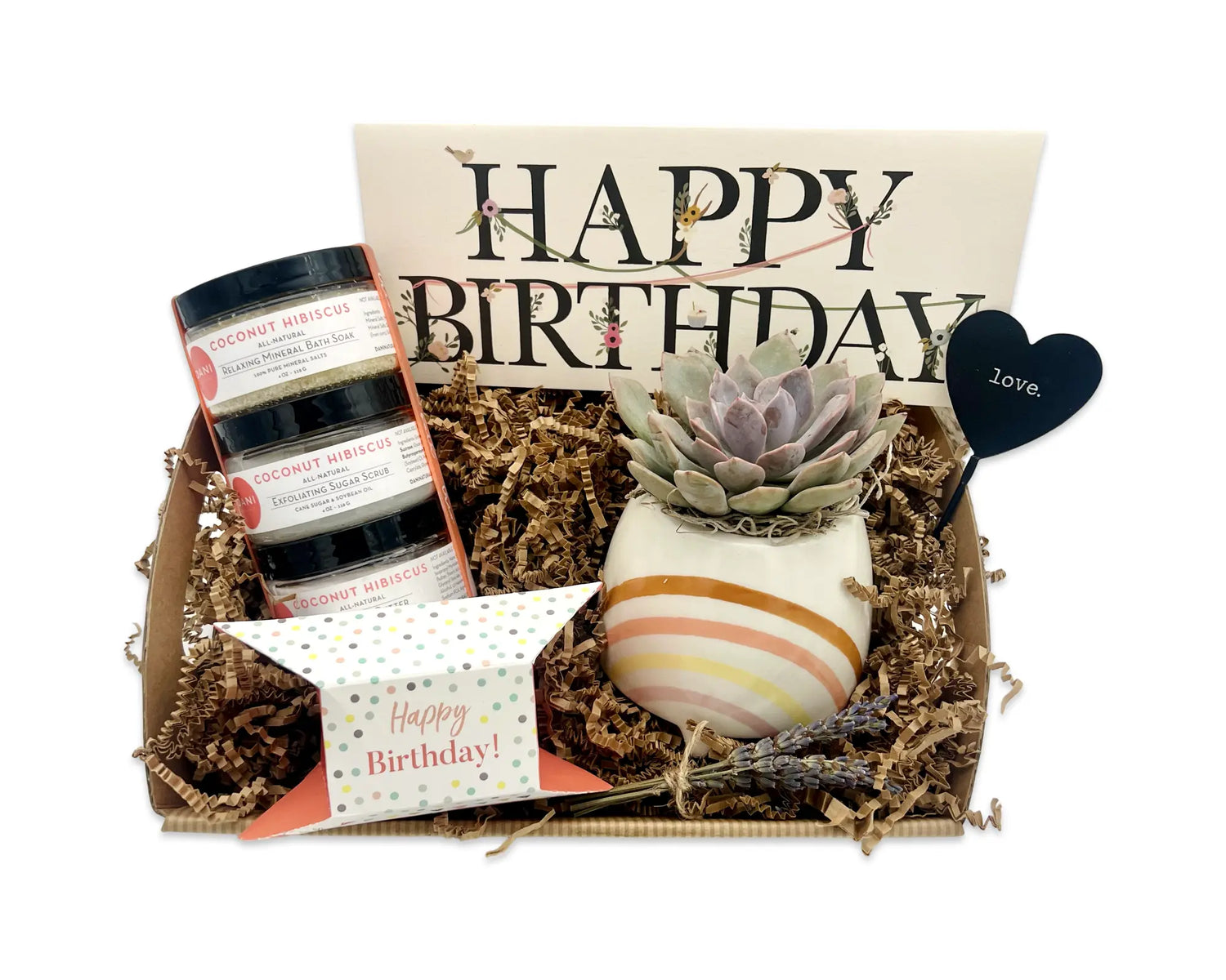 A gift set containing an assortment of bath and body products, including a shower steamer, mineral bath soak, sugar scrub, and body butter, all in coconut hibiscus scent, and a live succulent plant in a ceramic pot with a metal heart, wrapped in a signature box with 'Happy Birthday' text and dried lavender.
