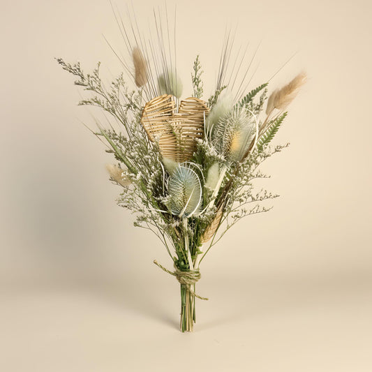 A dried floral arrangement featuring a natural woven heart, thistles, pampas grass, and delicate filler stems, tied with jute twine at the base, set against a solid warm beige background.
