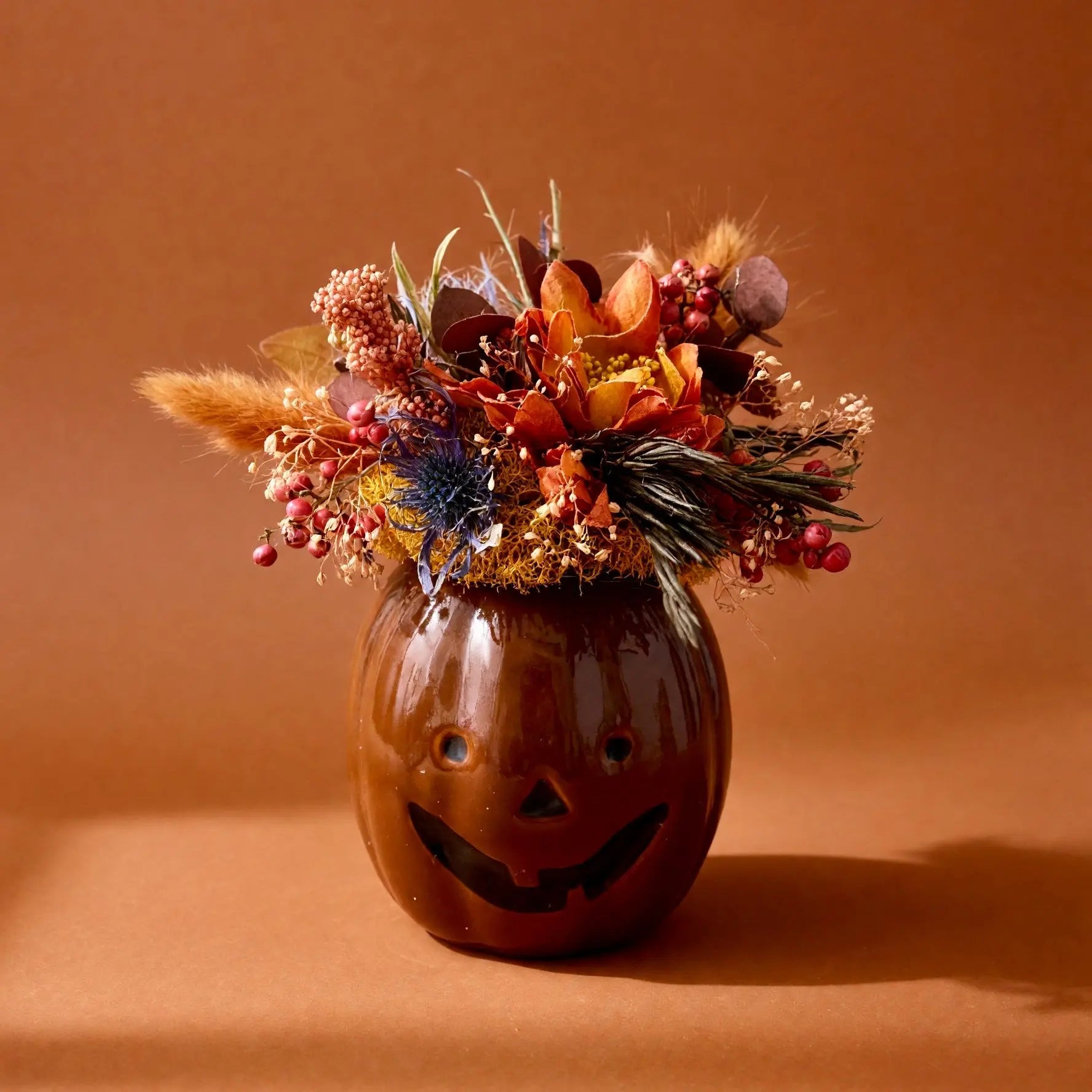 A decorative pumpkin-shaped vase with a carved jack-o'-lantern face, filled with an autumn floral arrangement of dried grasses, berries, and small orange flowers, set against a warm brown studio background.