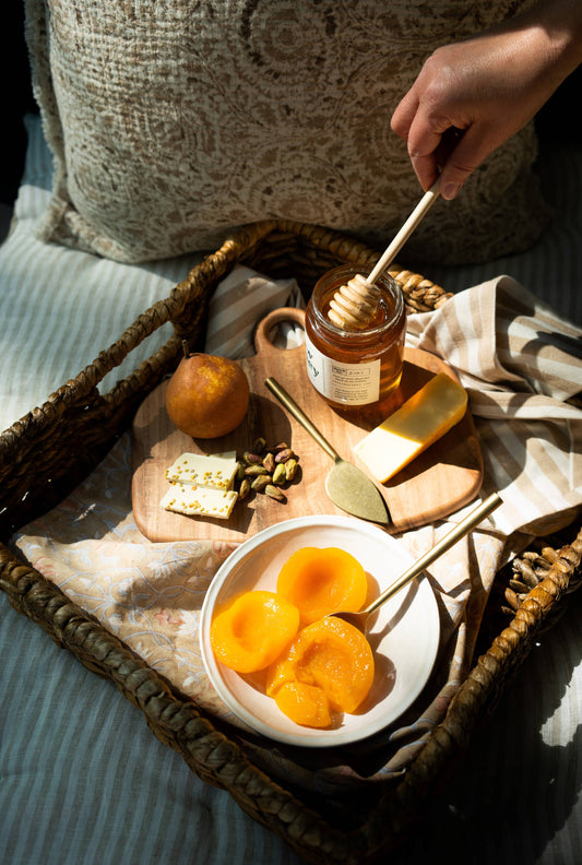 display of fruit and honey with cheese knives and wooden cutting board in a tray on a bed
