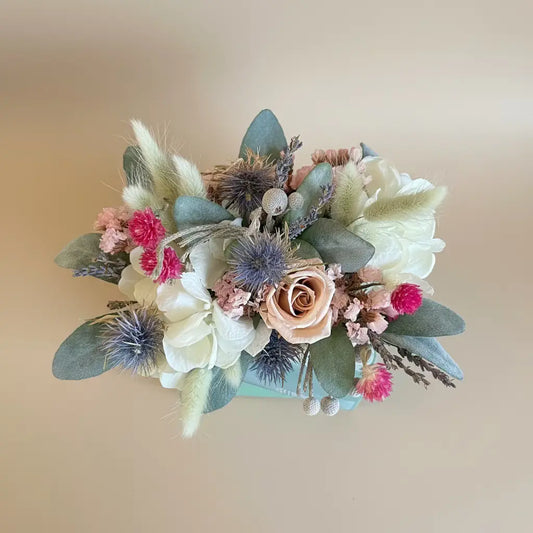 A dried floral arrangement featuring a peach rose, white flowers, pink pom-pom blossoms, and muted blue-green foliage on a dark reflective surface.