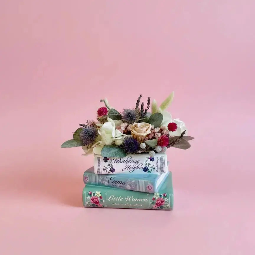 A decorative arrangement of three stacked vintage-style books in teal, white, and pink floral detailing, topped with a small bouquet of faux flowers, set against a solid pink background.