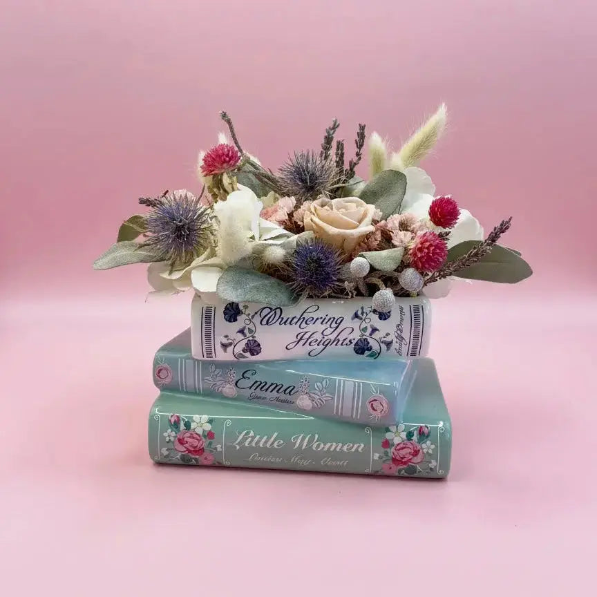 A decorative floral arrangement placed on top of three stacked books with titles including 'The Wuthering Heights', 'Emma', and 'Little Women'. The flowers include cream roses, purple thistle-like blooms, red berries, and green foliage, set against a soft pink studio background.