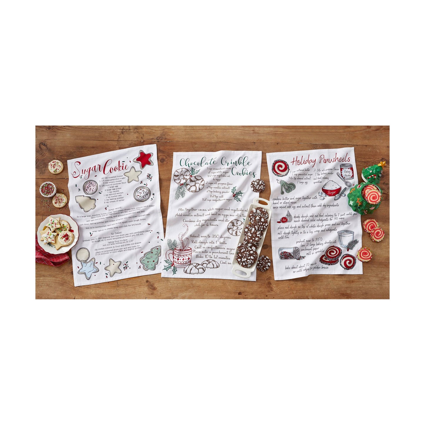 Three recipe dishtowels for sugar cookie, chocolate crinkle cookies, and holiday cookies on a wooden surface with decorative elements.