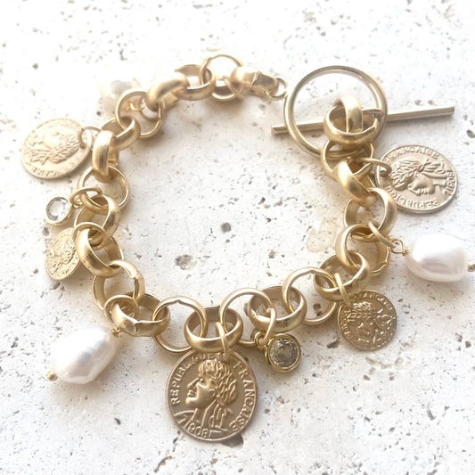 Gold charm bracelet with coins and pearls on a textured white surface