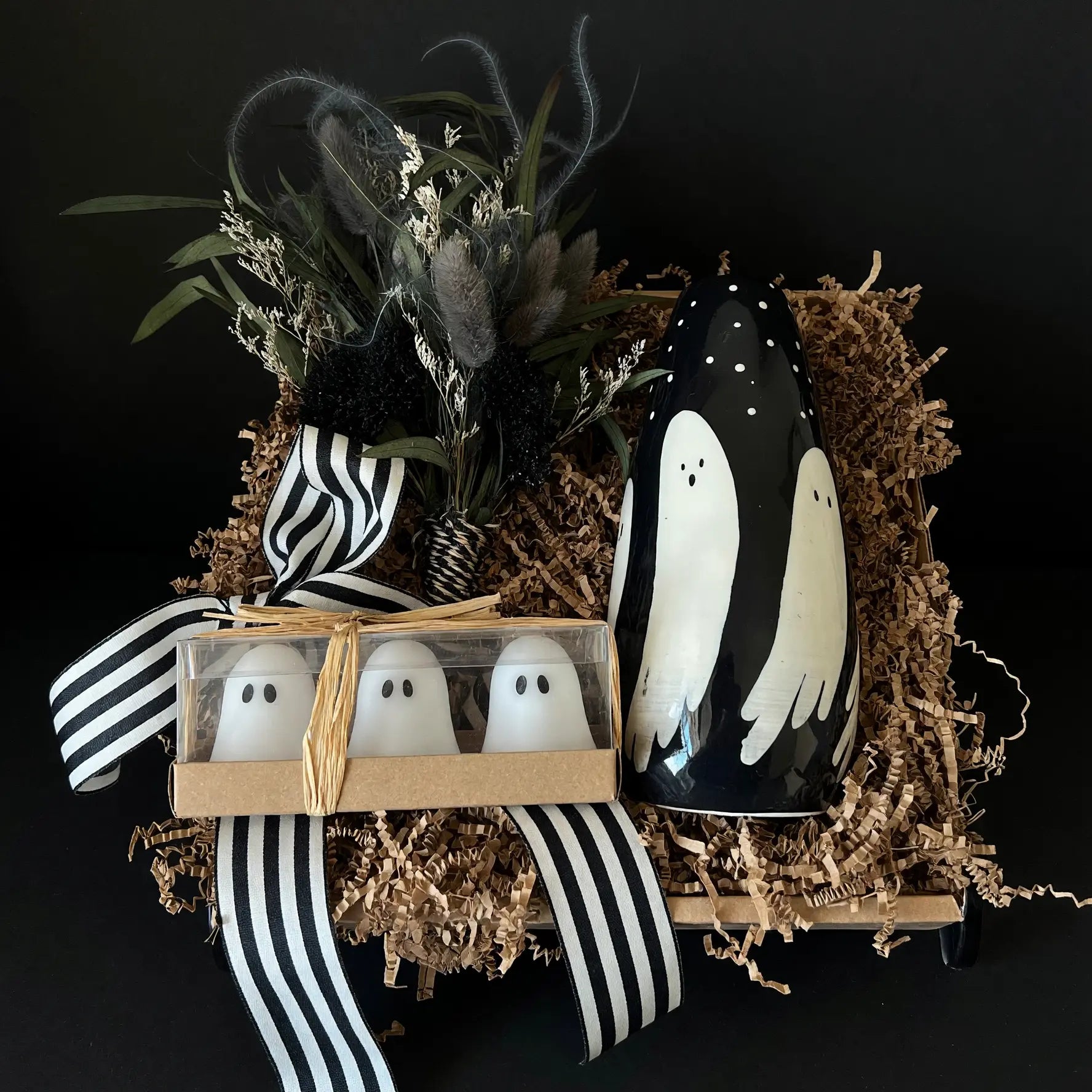 Halloween-themed gift set featuring a black ceramic vase with white ghost illustrations, a small box of three white ghost figurines in clear packaging, dried botanicals, and black-and-white striped ribbon, all arranged in brown shredded paper against a black background.