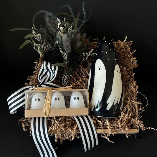 Halloween-themed gift set featuring a black ceramic vase with white ghost illustrations, a small box of three white ghost figurines in clear packaging, dried botanicals, and black-and-white striped ribbon, all arranged in brown shredded paper against a black background.