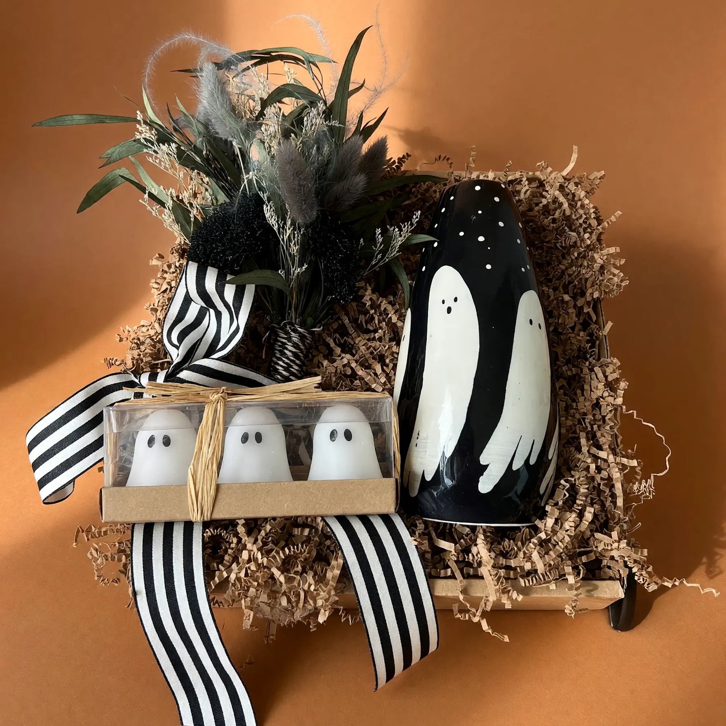 A Halloween-themed gift arrangement featuring a black ceramic ghost-shaped candle, a small box of three white ghost-shaped salt and pepper shakers, and dried botanicals tied with a black-and-white striped ribbon, all set in brown paper shred against a warm terracotta background.