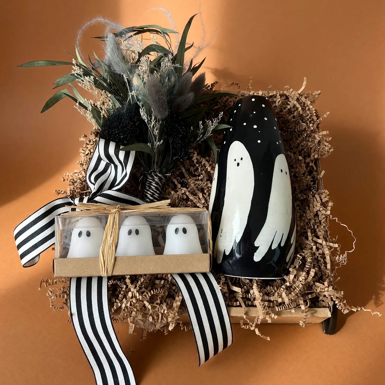 A Halloween-themed gift arrangement featuring a black ceramic ghost-shaped candle, a small box of three white ghost-shaped salt and pepper shakers, and dried botanicals tied with a black-and-white striped ribbon, all set in brown paper shred against a warm terracotta background.