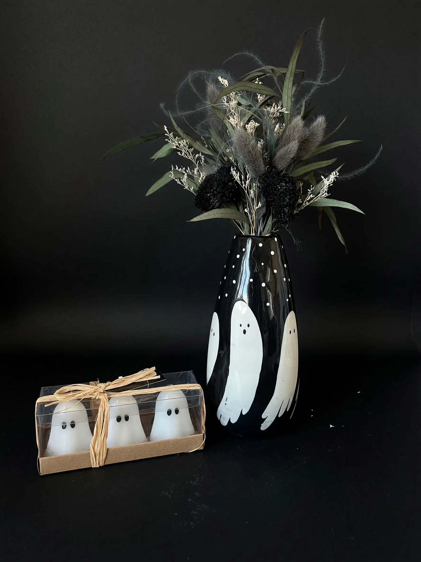 A black ceramic vase decorated with white ghost illustrations, filled with dried green and black botanicals, standing next to a small clear box containing three matching white ghost figurines tied with a tan ribbon, all set against a dark black background.