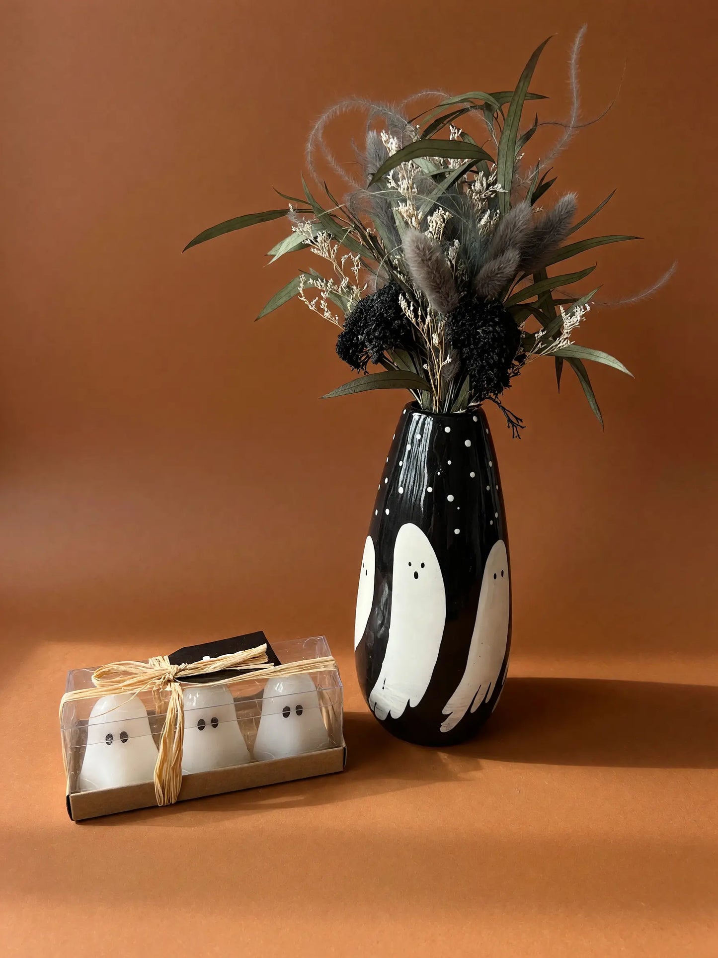 A black ceramic vase with white ghost illustrations and dried floral arrangements, shown next to a small gift box containing two white ghost-shaped items tied with twine, on a warm brown studio background.