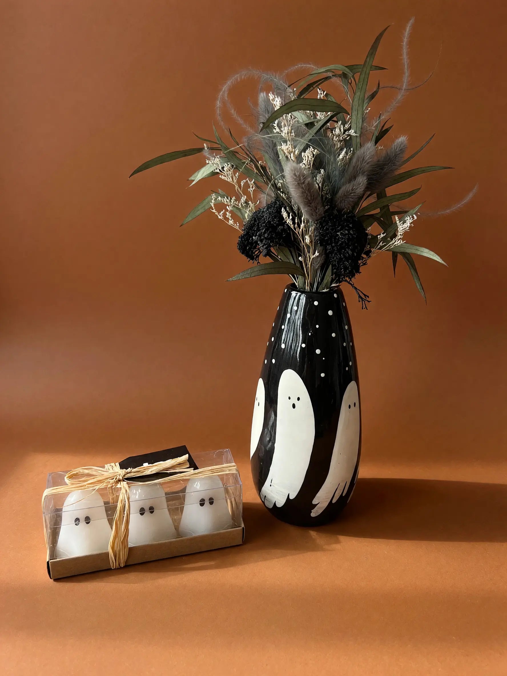 A black ceramic vase with white ghost illustrations and dried floral arrangements, shown next to a small gift box containing two white ghost-shaped items tied with twine, on a warm brown studio background.
