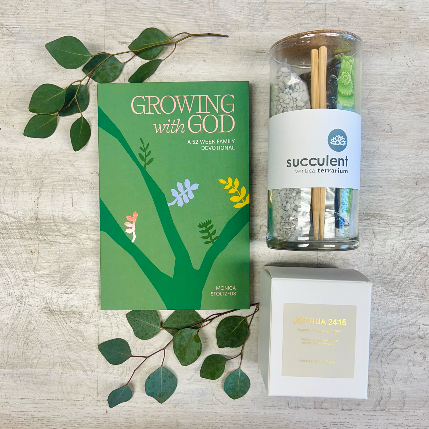 A gift set including a succulent terrarium kit, a family devotional book titled 'Growing with God', and a candle with a label that reads 'Joshua 24:15'. All items are displayed on a wooden surface with decorative green leaves.