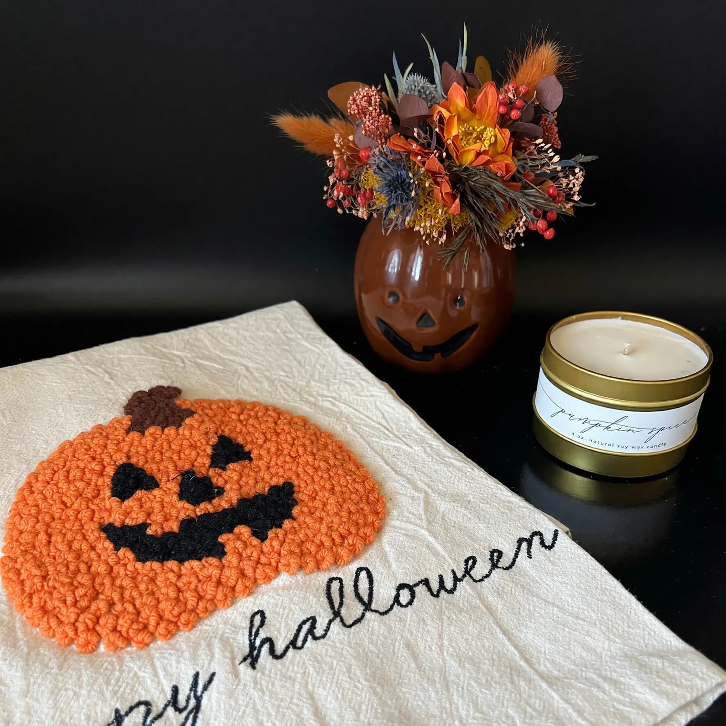 A Halloween-themed decorative scene on a black surface, featuring a cream fabric cloth with an orange punch-needle jack-o'-lantern and the words 'Happy Halloween' in black script. Behind it is a small brown pumpkin-shaped vase filled with dried autumn flowers and a white candle in a gold tin.