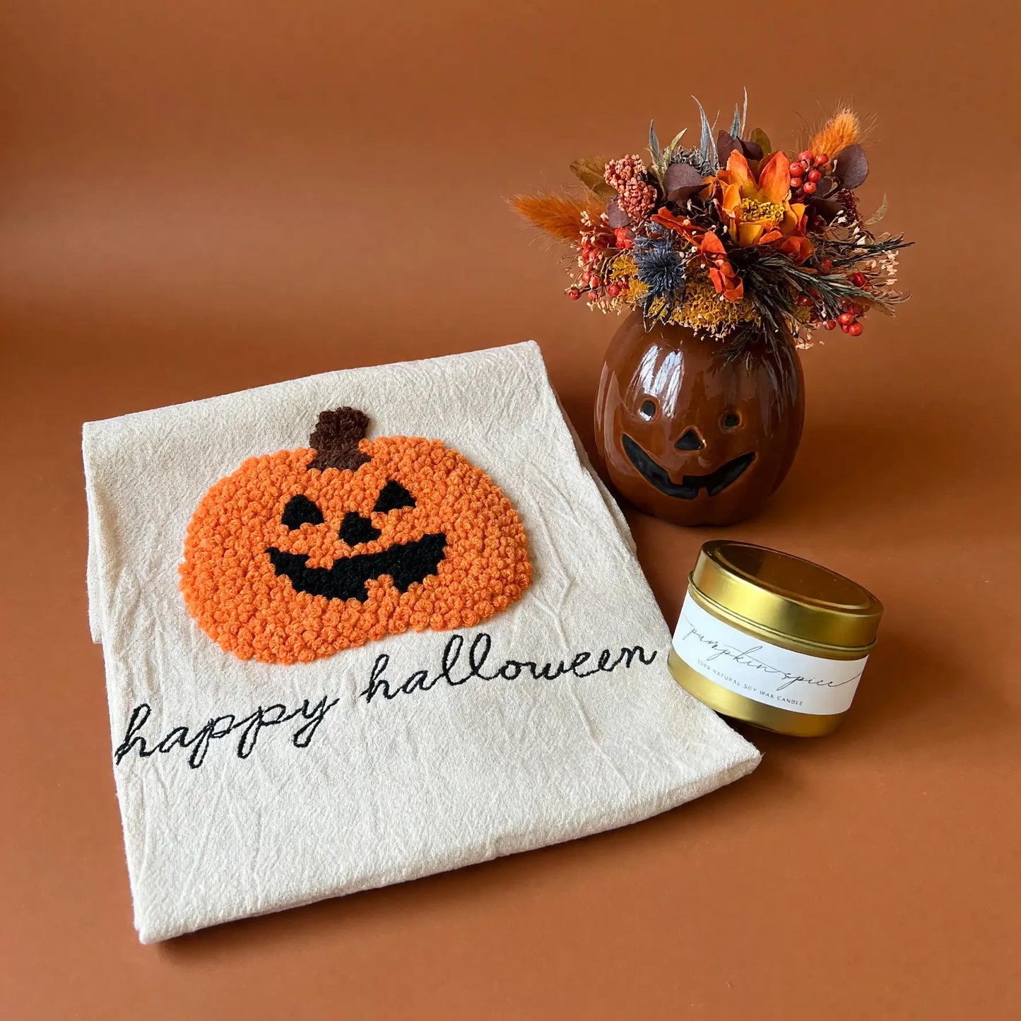 A styled flat-lay featuring a cream-colored halloween kitchen towel with a textured orange and black pumpkin design and the words 'happy halloween' in black script. Next to it is a small brown glass jar with a carved jack-o'-lantern face and a gold lid, filled with autumn-colored dried flowers, all set against a solid terracotta background.