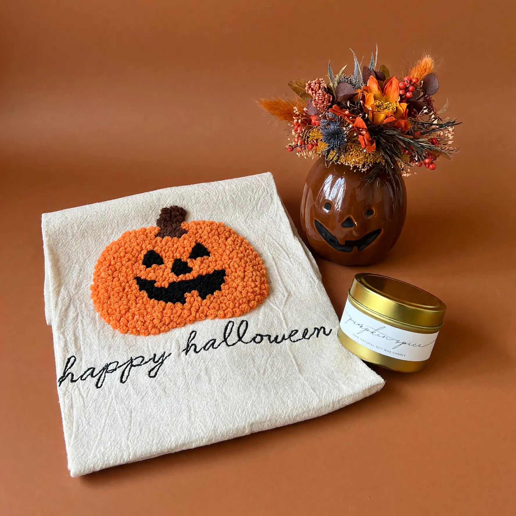 A styled flat-lay featuring a cream-colored halloween kitchen towel with a textured orange and black pumpkin design and the words 'happy halloween' in black script. Next to it is a small brown glass jar with a carved jack-o'-lantern face and a gold lid, filled with autumn-colored dried flowers, all set against a solid terracotta background.