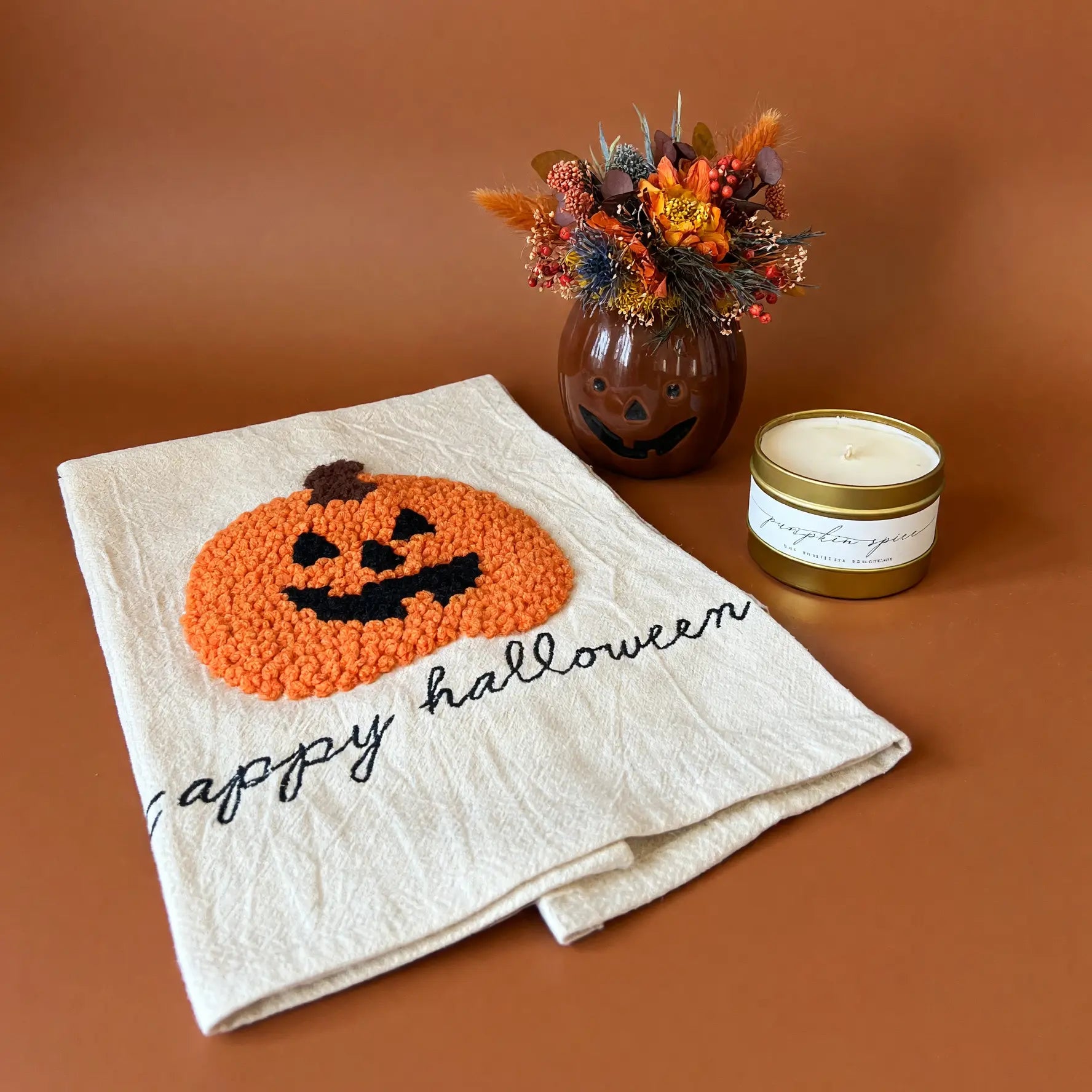 A folded cream-colored kitchen towel with a textured orange and black pumpkin design and the words 'happy halloween' embroidered in black script. The towel is styled on a warm brown background next to a small decorative vase with autumn foliage and a candle.