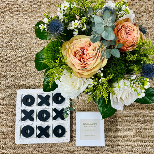 A curated home gift set featuring a black and white terrazzo tic-tac-toe game set, a soy candle, and a fresh flower arrangement in a glass vessel, all displayed on a neutral background.