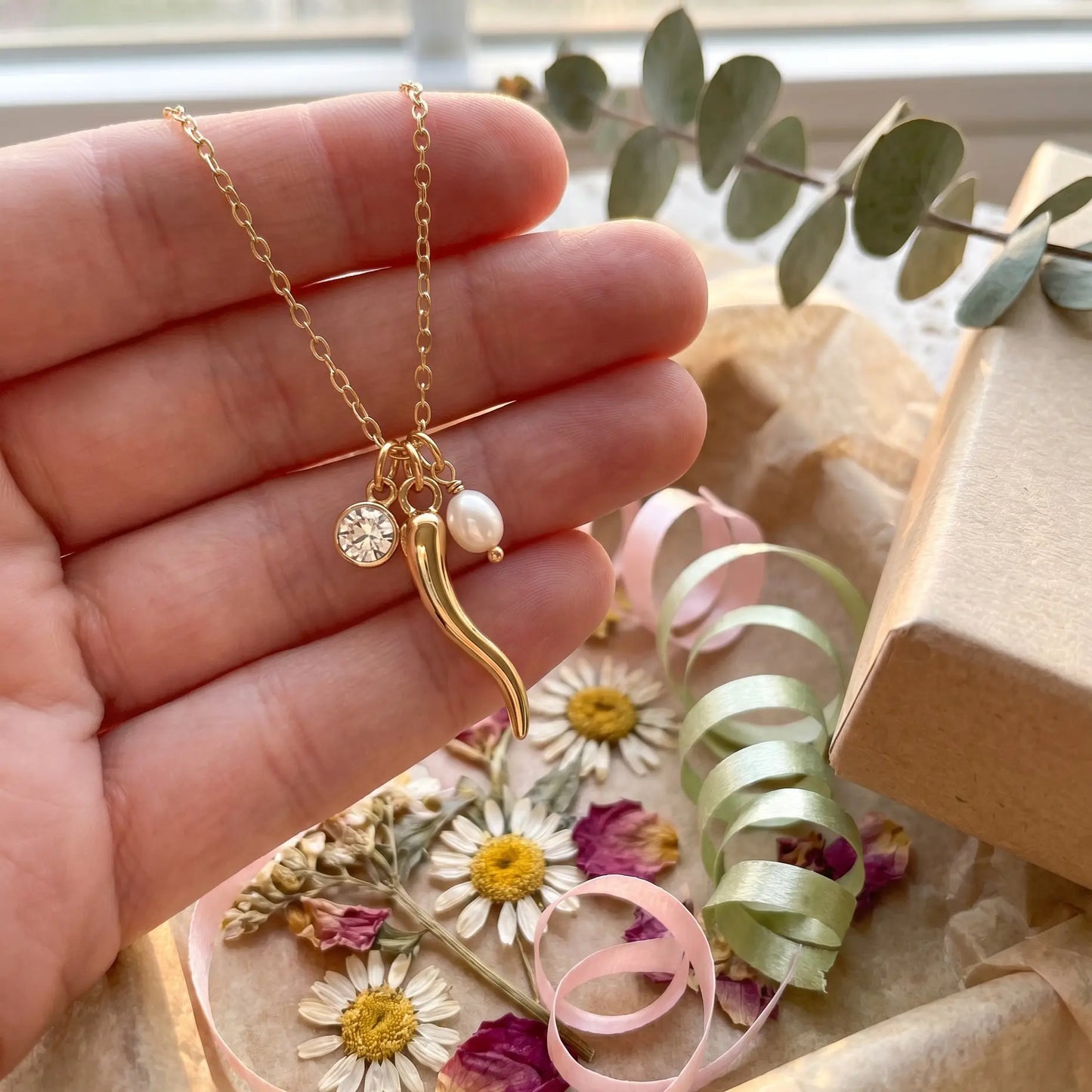 Gold necklace with a pendant held by a hand, with flowers and ribbons in the background.