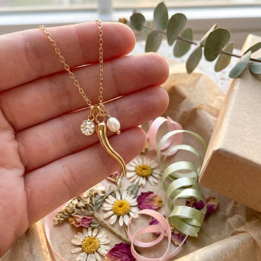 Gold necklace with a pendant held by a hand, with flowers and ribbons in the background.
