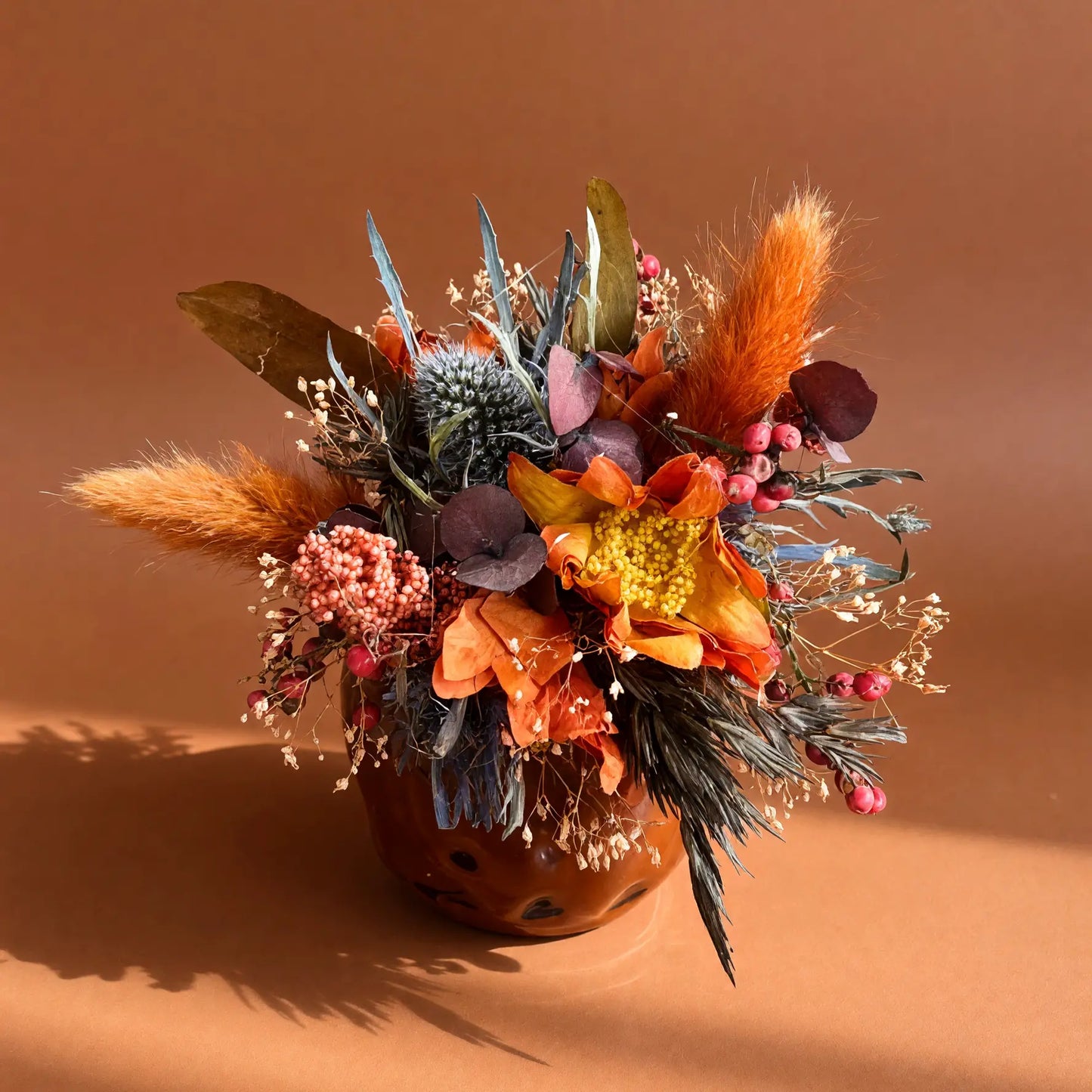 A dried floral arrangement in a terracotta-colored pot, featuring orange flowers, blue-green foliage, and fluffy brown grasses, set against a solid warm brown background.