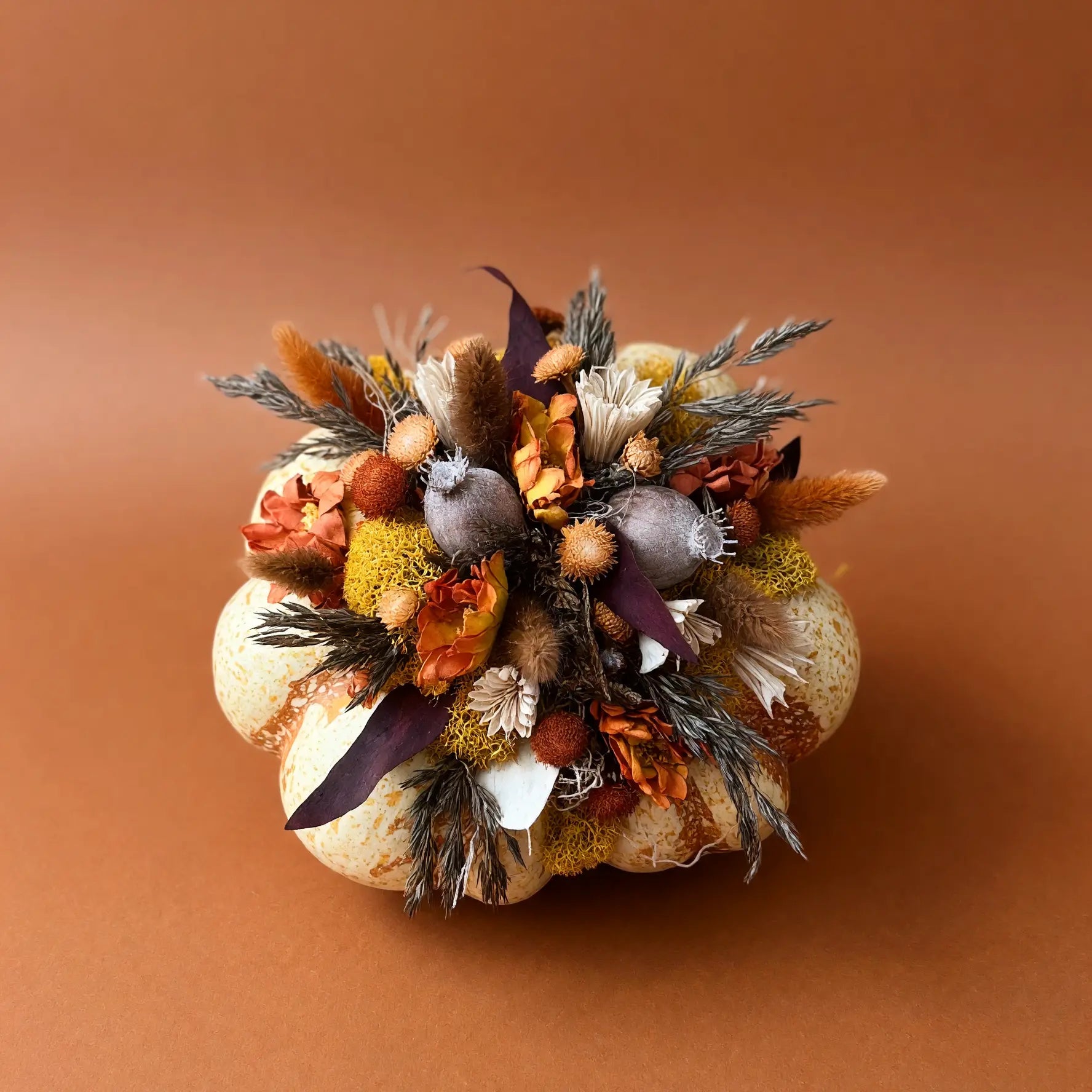 A decorative dried floral arrangement in a rounded, pumpkin-like base, featuring autumnal elements such as orange and brown grasses, beige and white flowers, and muted purple accents, photographed against a solid warm brown background.