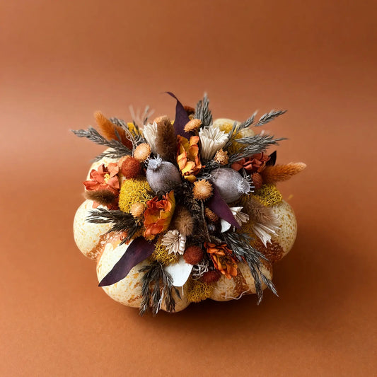 A decorative dried floral arrangement in a rounded, pumpkin-like base, featuring autumnal elements such as orange and brown grasses, beige and white flowers, and muted purple accents, photographed against a solid warm brown background.