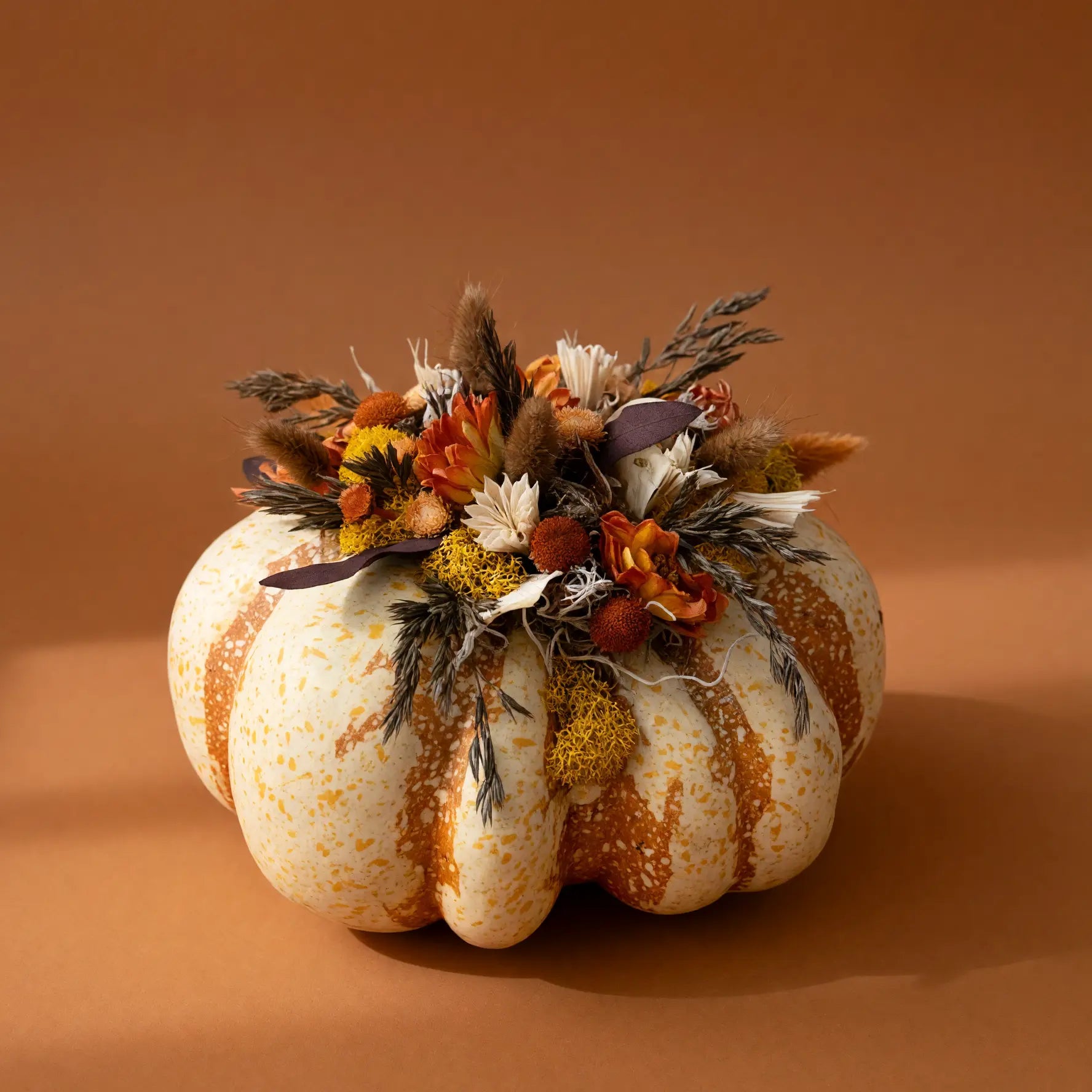 A decorative pumpkin in off-white with orange speckles, topped with a dried floral arrangement in autumn colors, set against a solid warm brown background.
