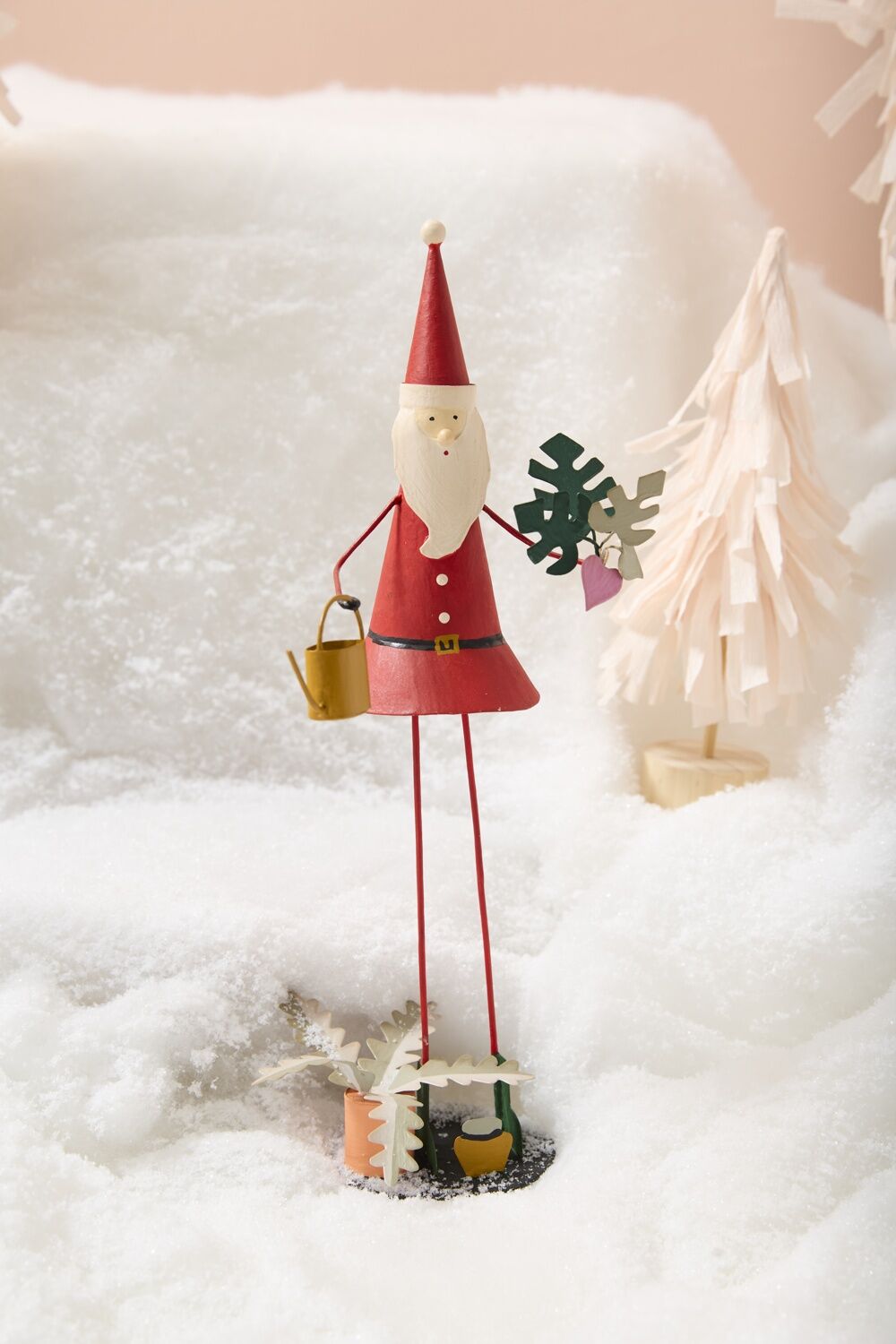 metal santa figurine with plants in snow with a light pink background