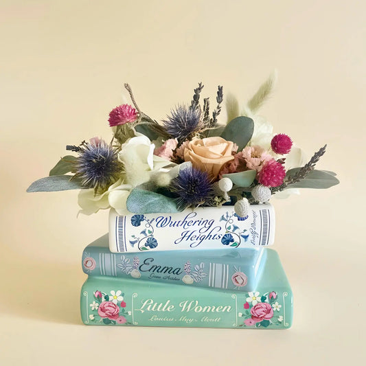 A decorative floral arrangement made from stacked books with floral covers, holding a bouquet of dried flowers including blue thistle, pink roses, and white blossoms, photographed against a plain warm beige background.