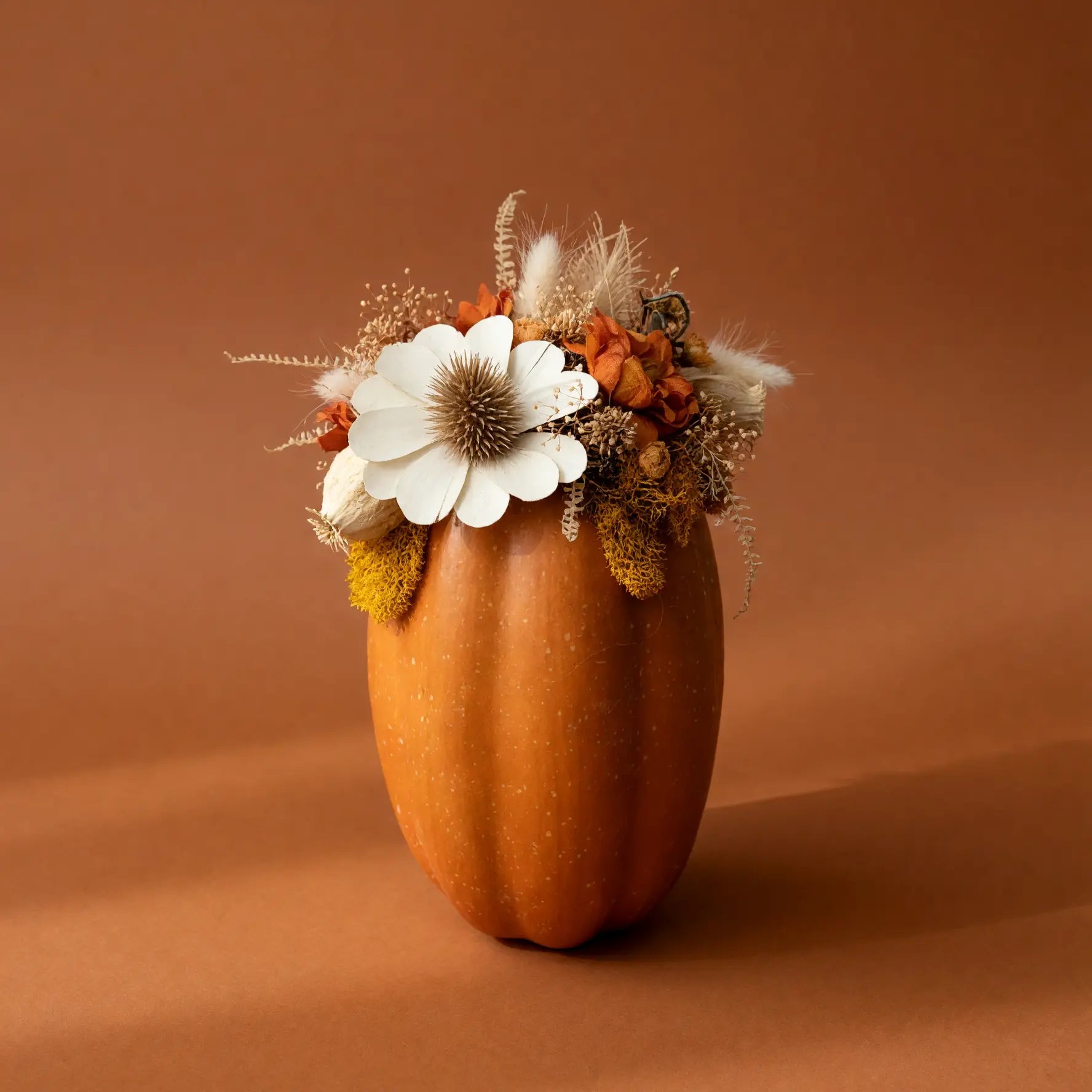 An orange decorative pumpkin with dried autumn flowers and foliage, set against a solid warm brown background.
