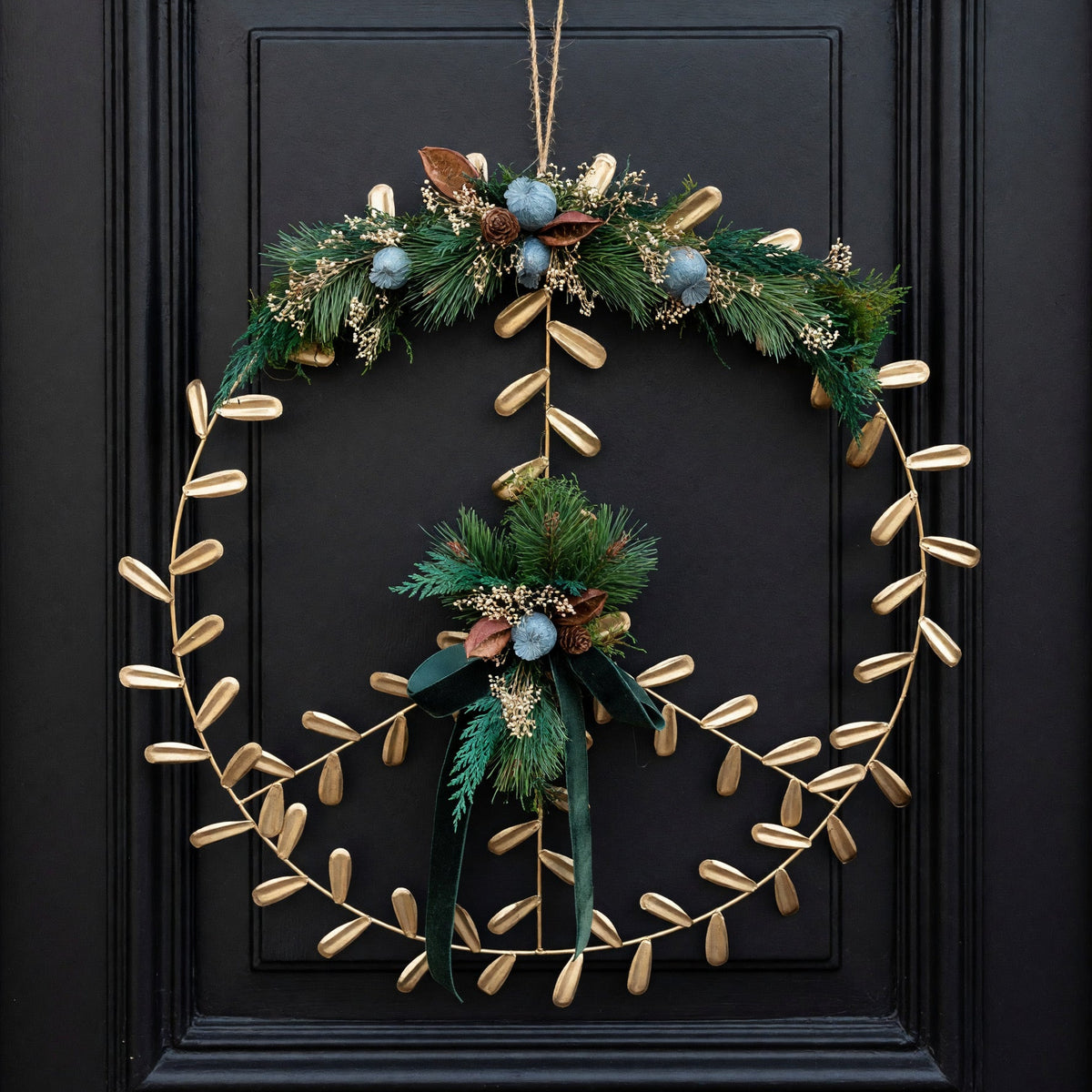 A decorative holiday wreath hanging on a black paneled door. The wreath features a gold leaf vine border with green pine branches, blue berries, and small brown accents, plus a dark green ribbon bow at the bottom.