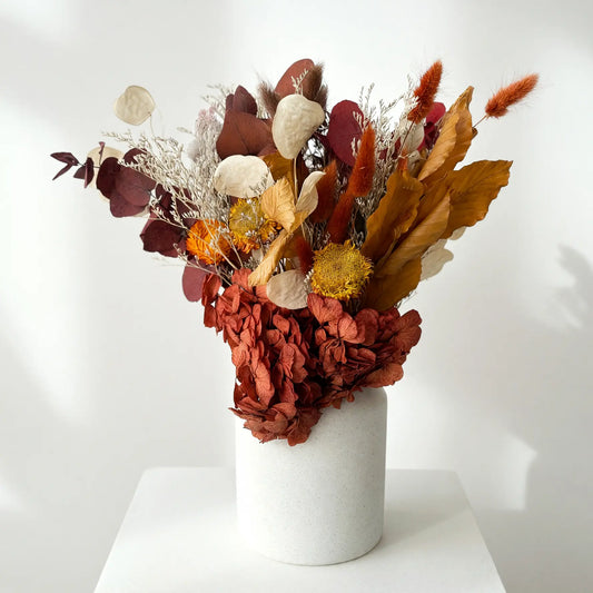 A dried flower arrangement in a matte white cylindrical vase, displayed on a white pedestal against a white wall with soft shadows.