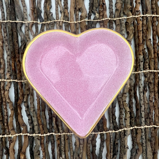 A heart-shaped porcelain trinket dish with a pink speckle glaze and hand-painted gold edge, displayed on a wicker surface.